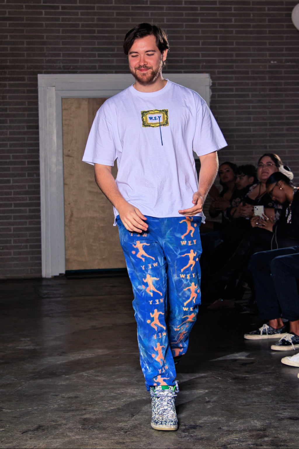 Person walking on a runway wearing a white t-shirt and blue pants with colorful patterns, with an audience in the background.