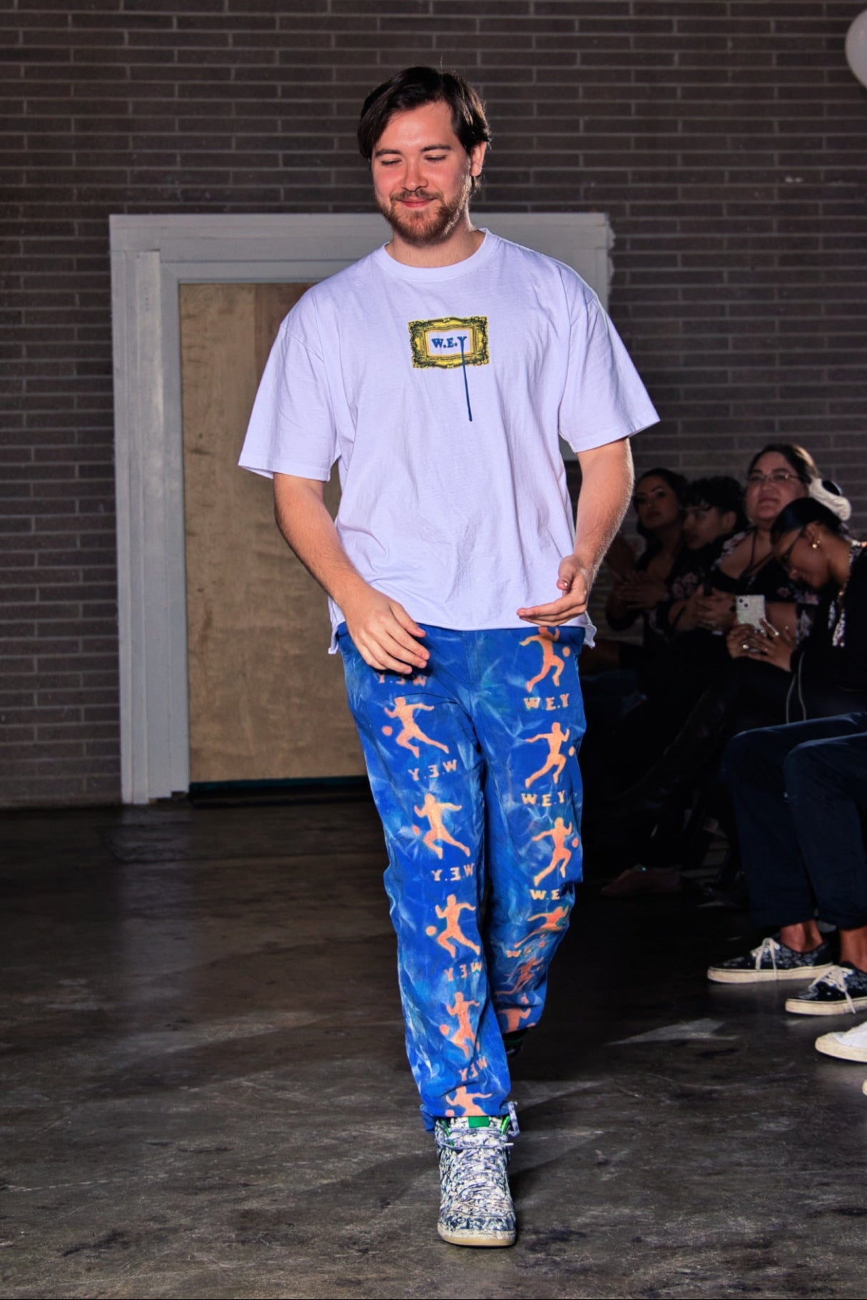 Person walking on a runway wearing a white t-shirt and blue pants with colorful patterns, with an audience in the background.