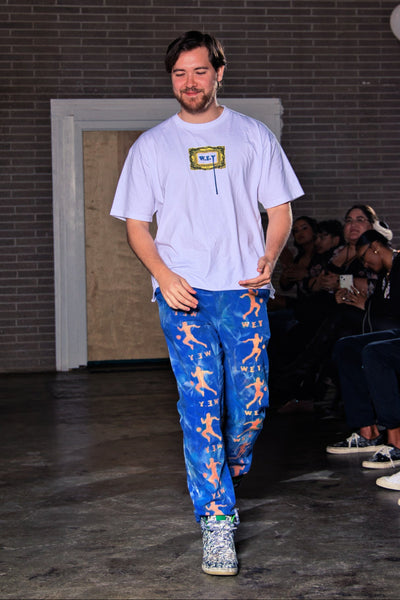 Person walking on a runway wearing a white t-shirt and blue pants with colorful patterns, with an audience in the background.
