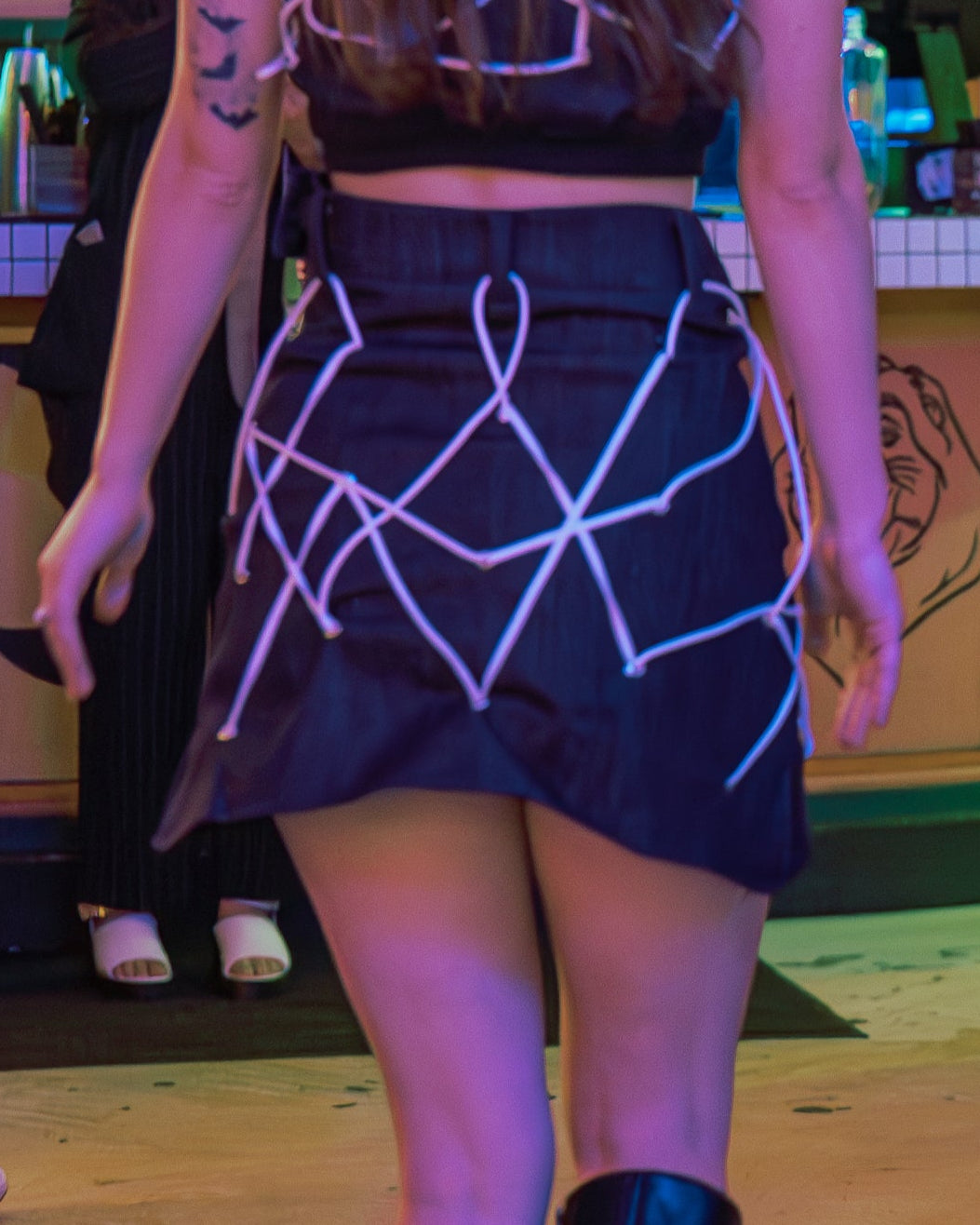 Two models walking on a runway in a fashion show with a bar and people in the background.